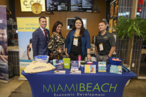 Four City of Miami Beach staff are standing behind a Miami Beach Economic Development booth at an event. The booth displays promotional materials, including brochures, pens, and branded items. A banner behind them reads "Say Hello to your new business," and the backdrop features a beach scene. The individuals are smiling and dressed professionally, with name badges visible. The setting appears to be indoors, at a networking or business expo. 