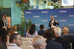 Miami Beach Mayor Steven Meiner speaker is addressing an audience at a Bisnow event, holding a microphone and gesturing while speaking. The backdrop features the Bisnow logo repeatedly on a blue banner. The audience is seated in white chairs, listening attentively. Behind the speaker, there are decorative plants, and the venue has vibrant green walls with a nature-themed design, including hanging plants and fish decorations. Two individuals stand on the left side, observing the event near a sign indicating restroom directions. The atmosphere suggests a professional, yet casual, networking or educational event.