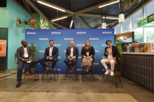 Five panelists are seated on high stools in front of a blue backdrop displaying the Bisnow logo, participating in a discussion at a professional event. Each panelist holds a microphone, engaging with the audience. The group includes three men in suits and two individuals in business attire, creating a balanced mix of perspectives. The venue has a casual and vibrant atmosphere, with colorful wall decorations featuring fish and plants. Behind the panel, a small food station is visible. The setting suggests a dynamic conversation on business, real estate, or economic development topics.