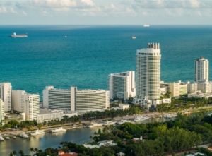 Aerial view of the Fontainebleau Hotel and surrounding Miami Beach skyline. The iconic curved hotel, along with adjacent high-rise buildings, sits along the beachfront, bordered by a waterway with yachts docked along its shore. The turquoise waters of the Atlantic Ocean stretch out to the horizon under a partly cloudy sky, with ships visible in the distance.