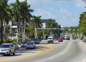 A sunny street view of 41st Street in Miami Beach, featuring cars driving in both directions, palm trees lining the sidewalks, and a mix of buildings in the background. The sky is clear and blue.